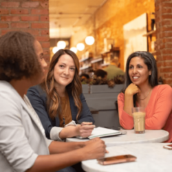 A man speaking with two women listening intently in a cafe together. They are dressed in business casual.